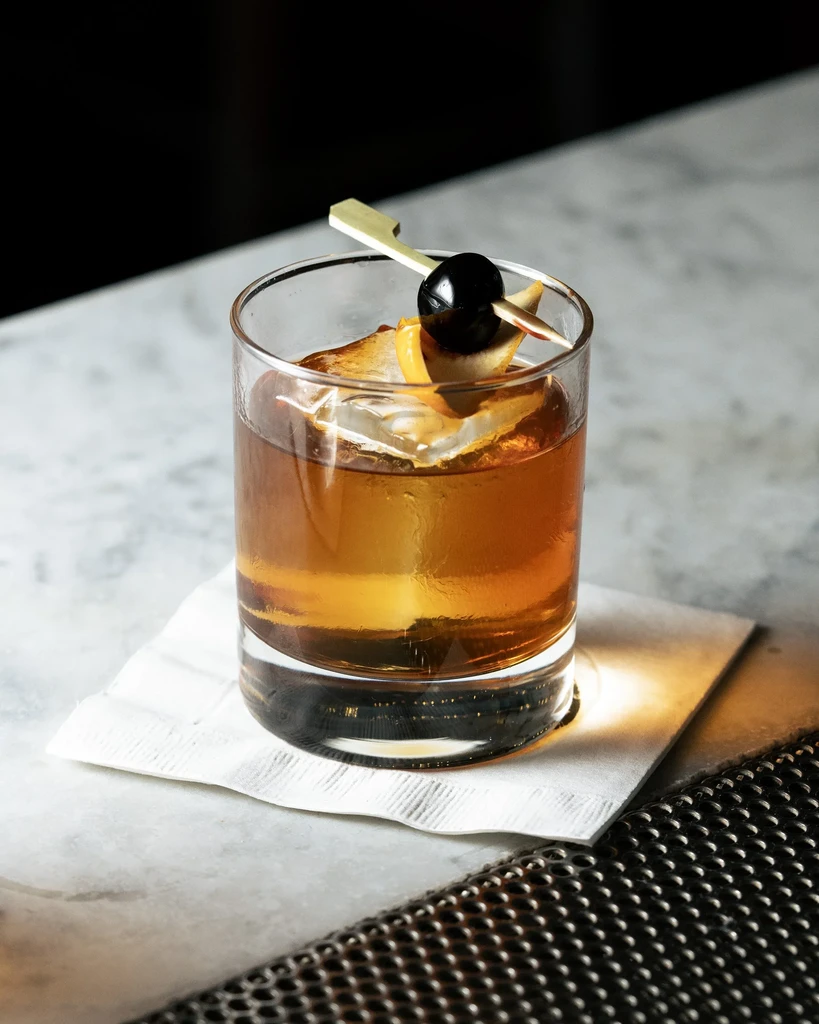 An amber cocktail with a cherry and orange peel on a bar.