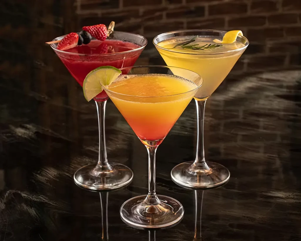 Three colorful cocktails in martini glasses on a dark reflective surface.