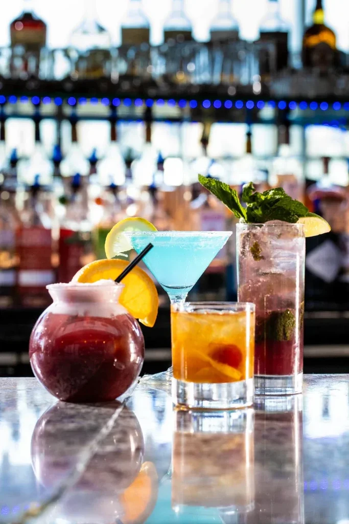 Four colorful cocktails on a bar with bottles in the background.