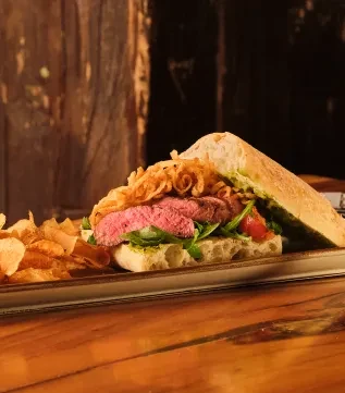 Steak sandwich with greens and fried onions, with chips on a wooden table.
