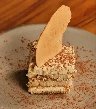 A rectangular piece of tiramisu with a decorative wafer and cocoa powder.
