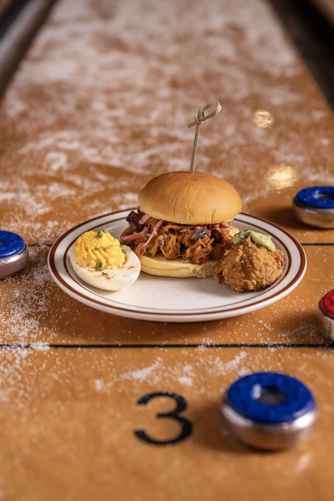 Plate with pulled pork slider, deviled egg, fried food on shuffleboard.