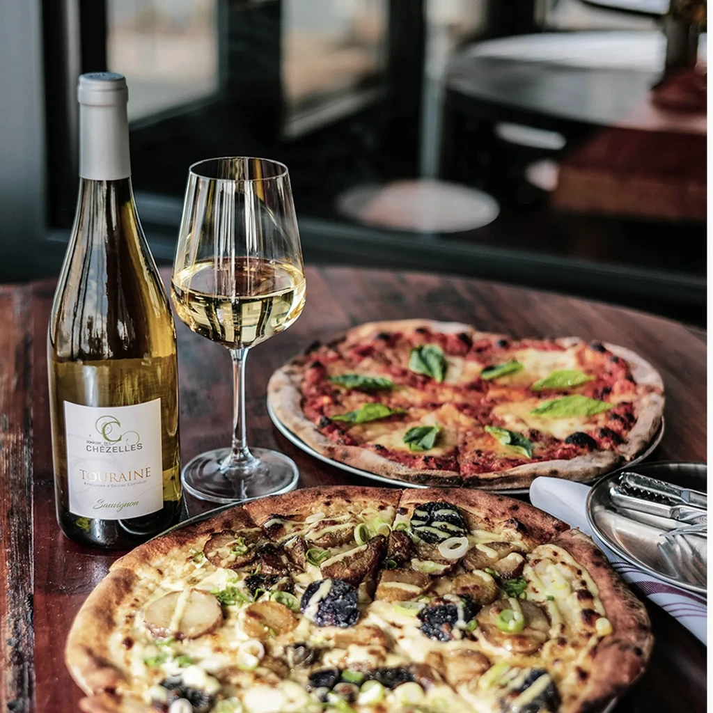Two pizzas, white wine bottle, and glass on a wooden table.