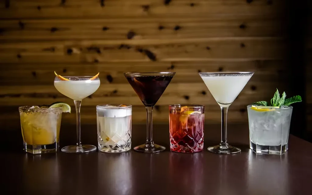 Six distinct cocktails on a bar counter with a wooden wall.