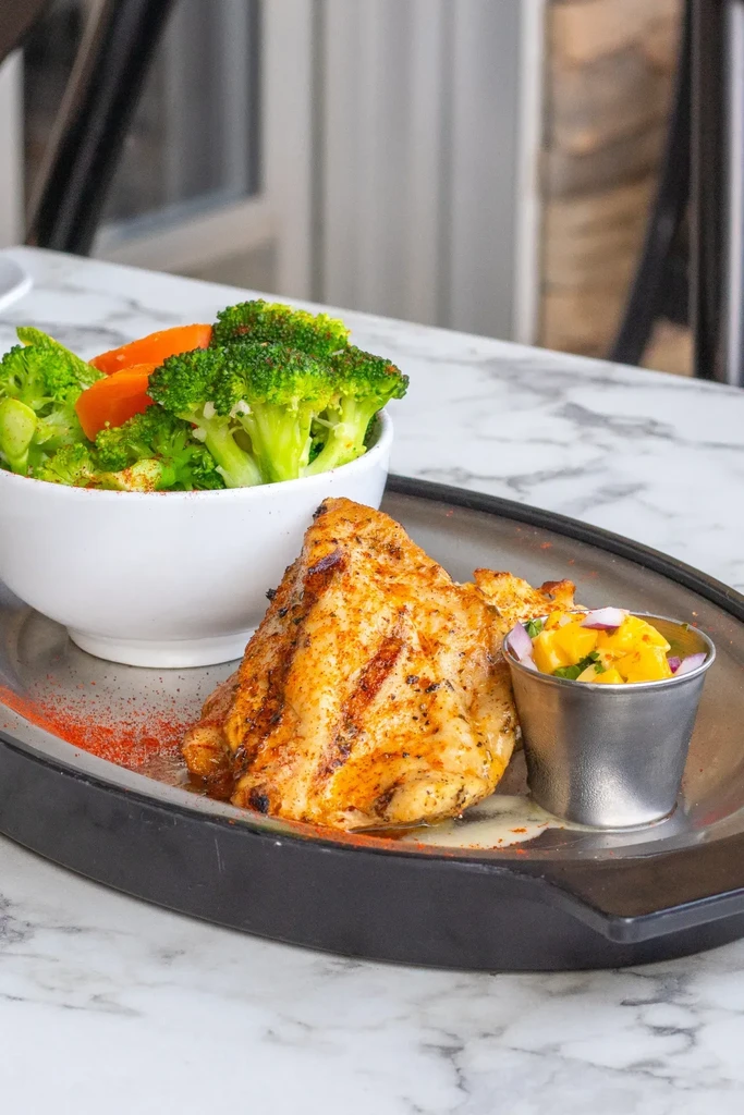 Grilled chicken with broccoli, carrots, and mango salsa on a marble table.