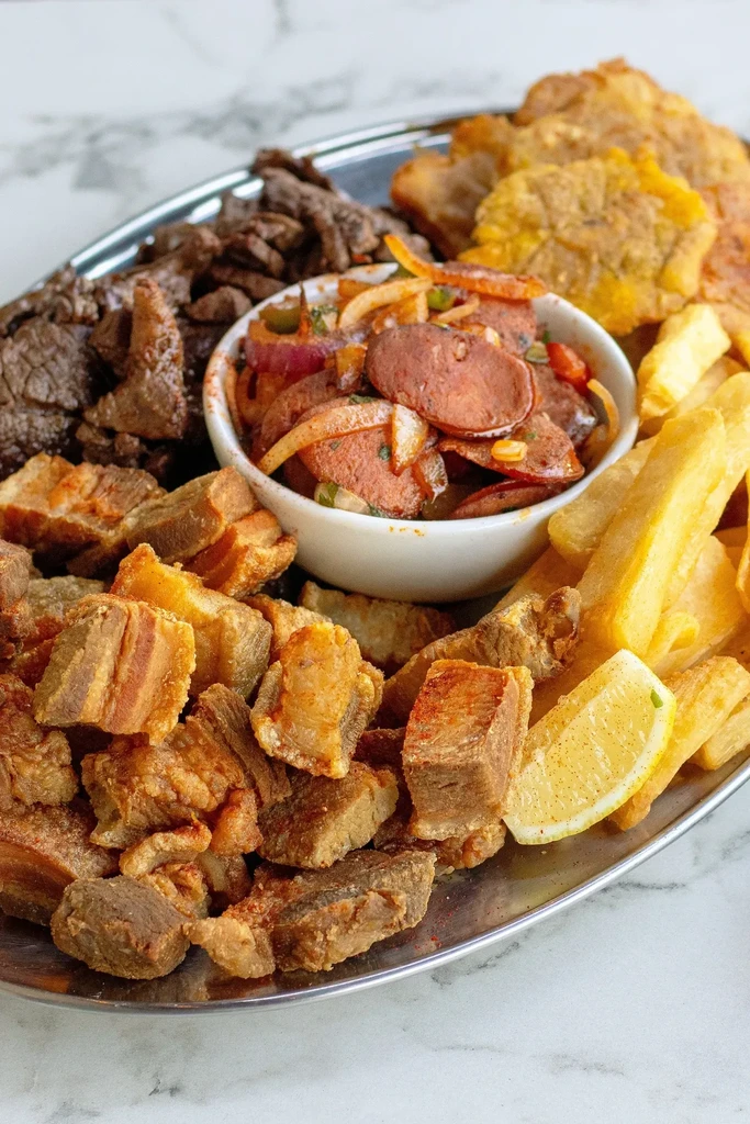 A platter of fried meat, sausage, plantains, and fries with a lemon wedge.