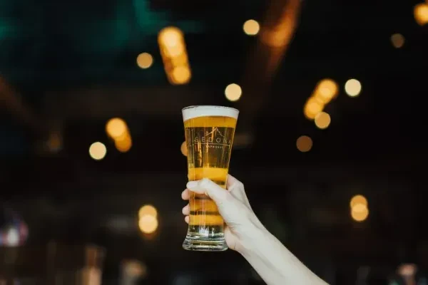 Hand holds Sedona glass of golden beer with frothy head, blurred lights.