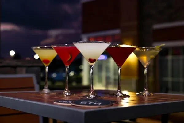 Five colorful cocktails in martini glasses on a table at dusk.
