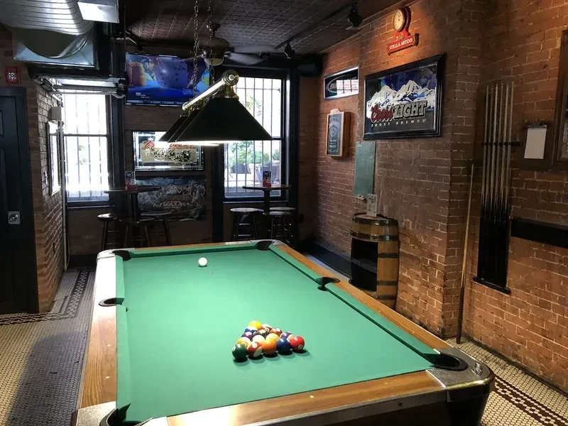 A pool table with balls racked, cue ball, in a brick-walled bar.