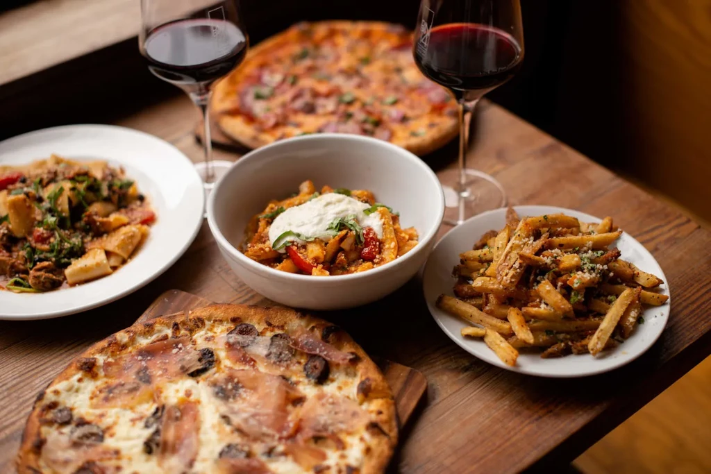 Two pizzas, two pasta dishes, and fries with two glasses of red wine.