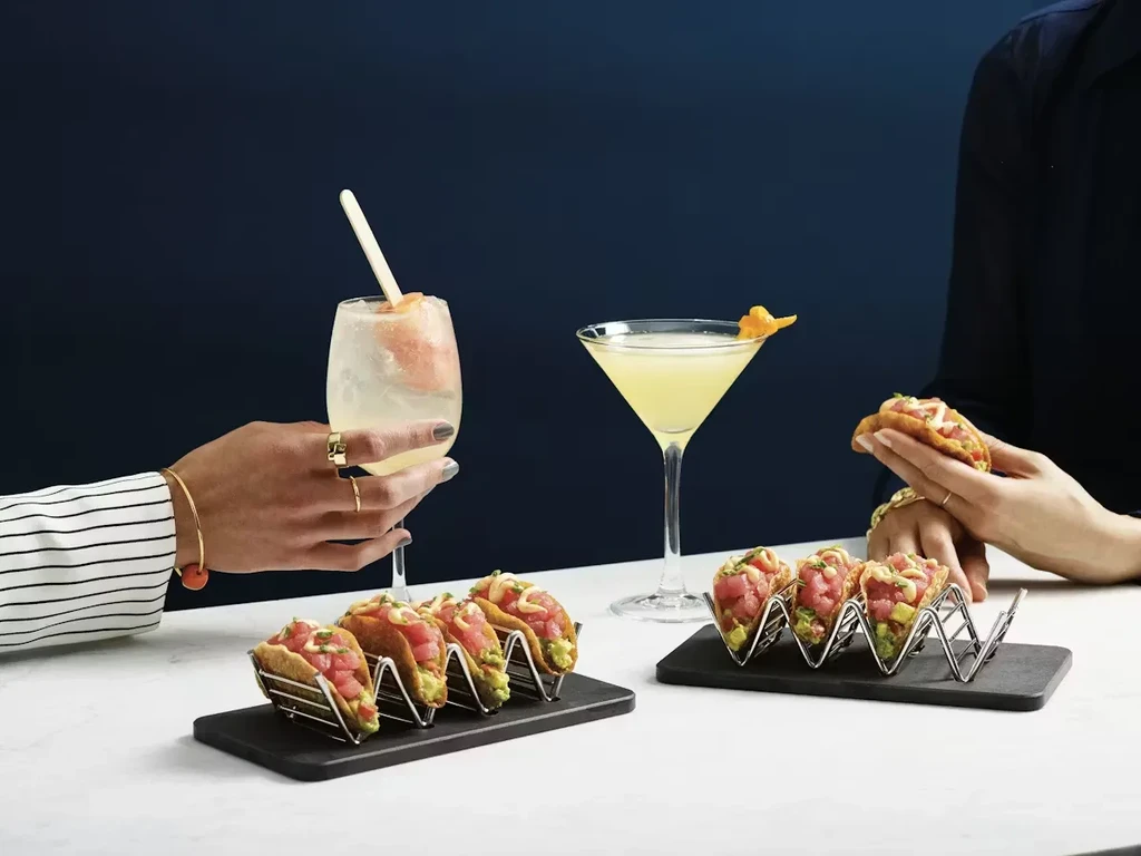 Two hands hold cocktails and mini tuna poke tacos on a table.