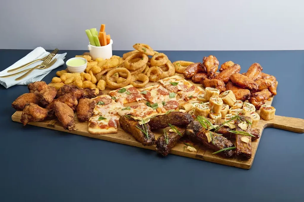 Wooden board filled with fried chicken, pizza, ribs, and fresh vegetables.
