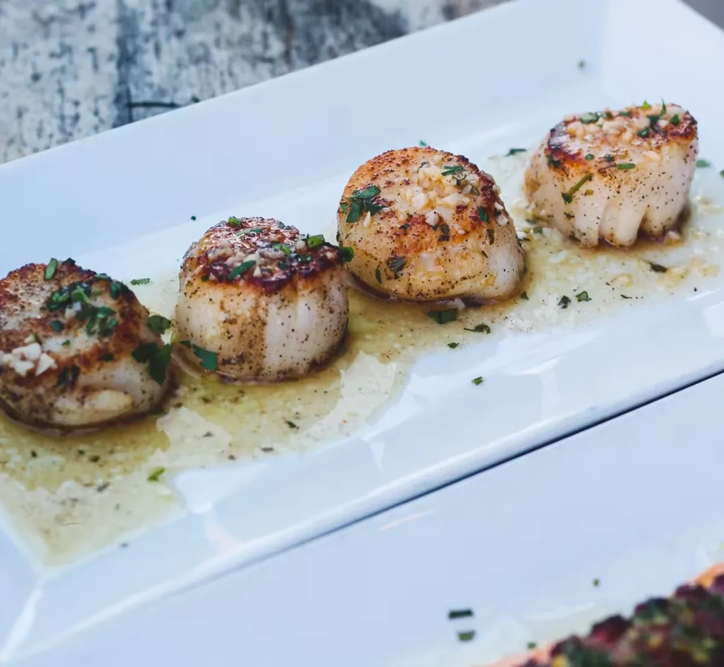 Four seared scallops with garlic butter and herbs on a white plate.