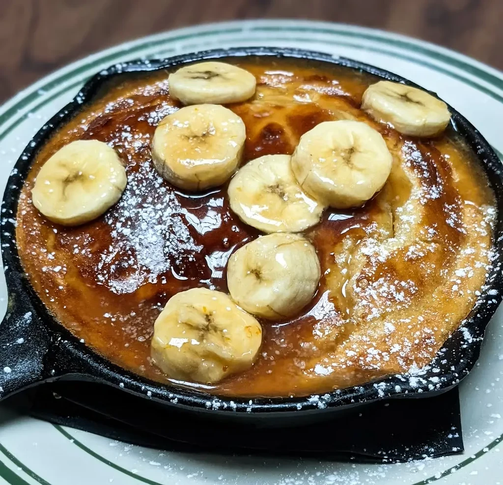A banana pancake skillet with powdered sugar and syrup, on a plate.