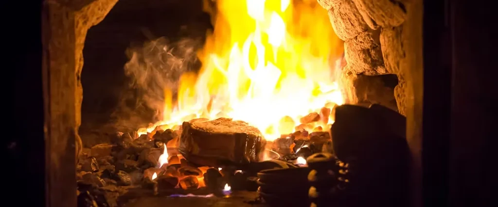A steak cooks on hot coals and a roaring fire in a hearth.