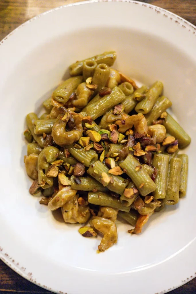 Green pasta with chopped pistachios and mushrooms in a white bowl.