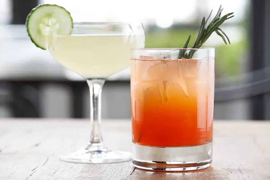 Two cocktails on a table: one pale with cucumber, one orange with rosemary.
