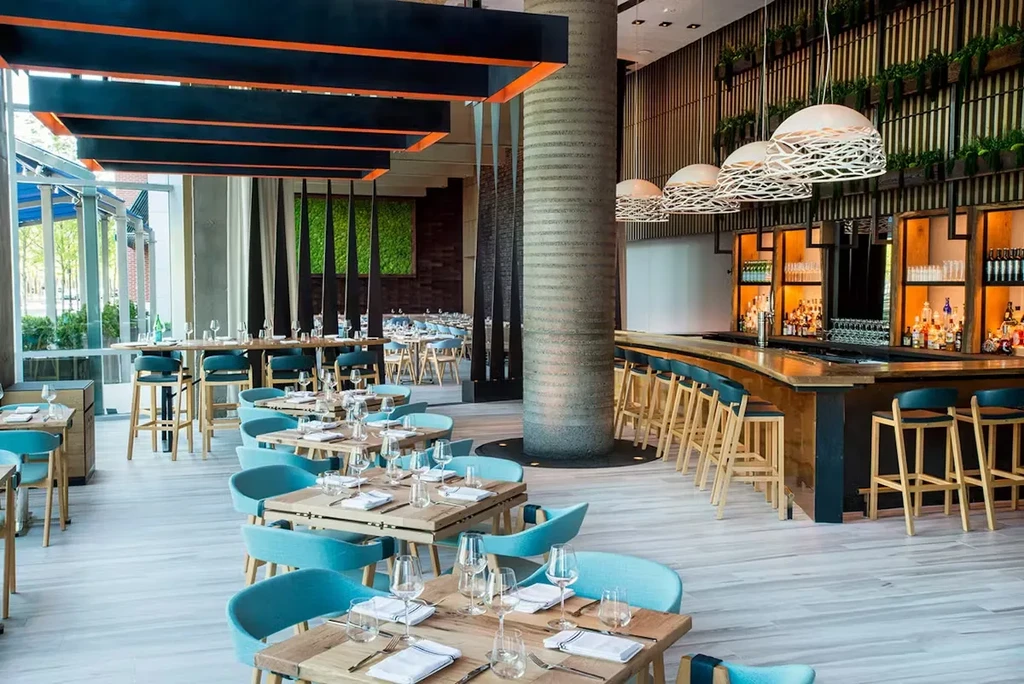 A modern restaurant interior with light wood tables, blue chairs, and a bar.