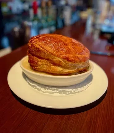A puffed pastry dome covers a white bowl of soup on a wooden table.