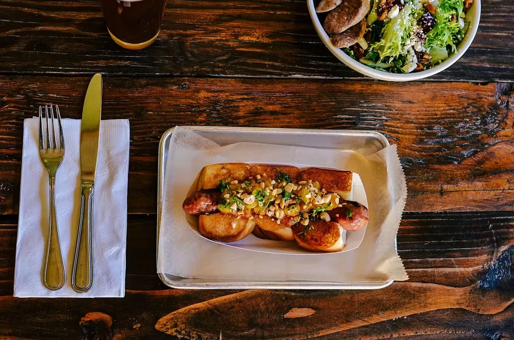 A hot dog with peanuts and herbs, a salad, and a drink on a wooden table.