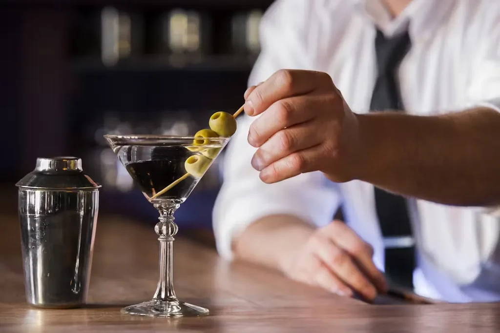 A bartender garnishes a martini glass with olives on a wooden bar.