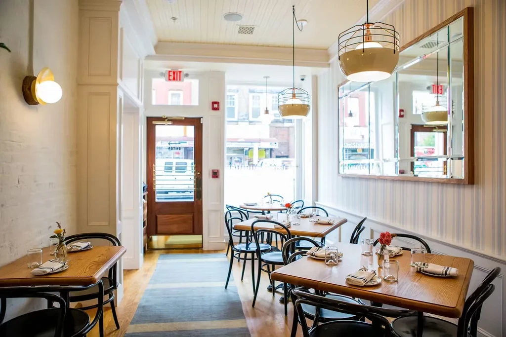 A bright, empty restaurant with wooden tables and black chairs.