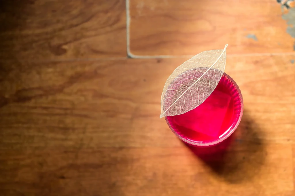 A skeleton leaf on a glass of red liquid on a wooden table.