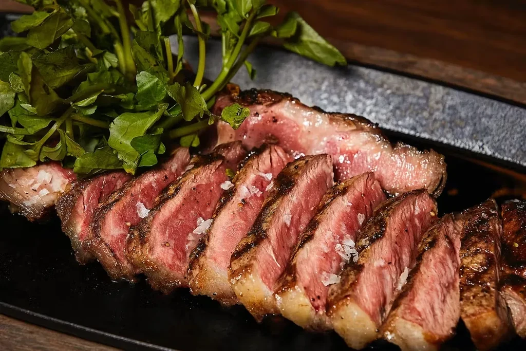 Sliced rare steak with coarse salt and green garnish on a black plate.