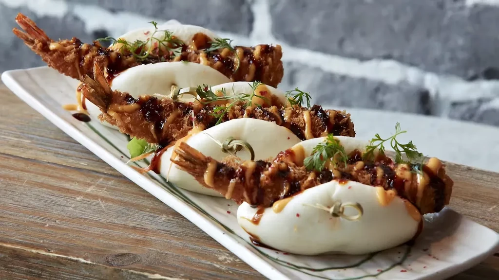 Four fried shrimp bao buns drizzled with sauces and herbs.