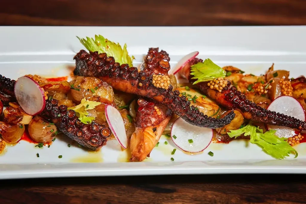 A rectangular white plate with roasted octopus tentacles, potatoes, radishes, and celery.