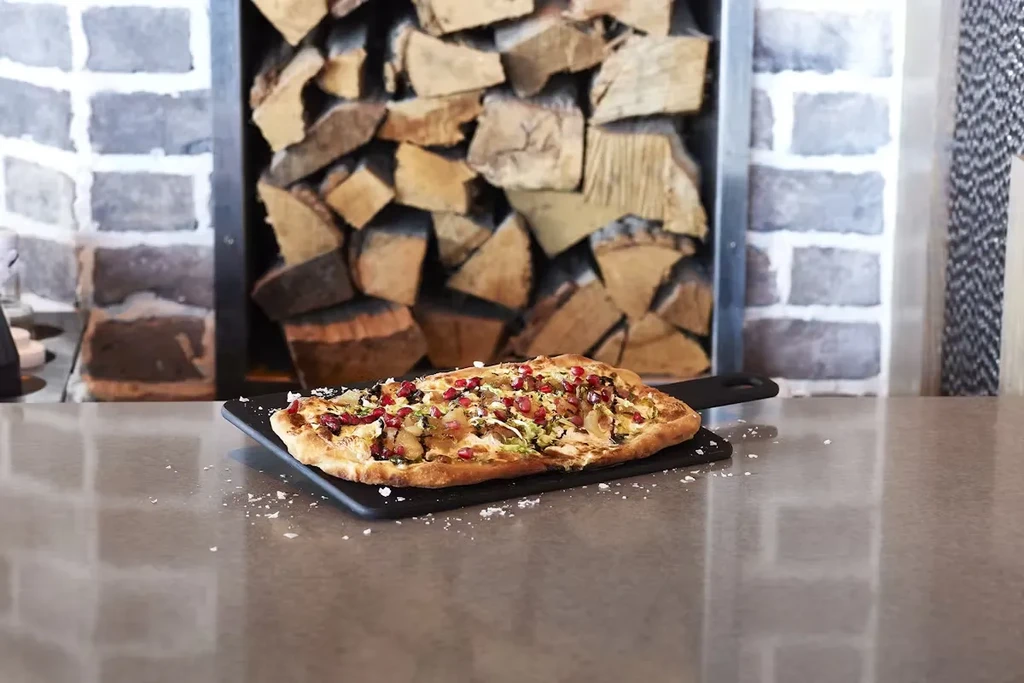 A flatbread pizza with pomegranate seeds on a table, with firewood in background.
