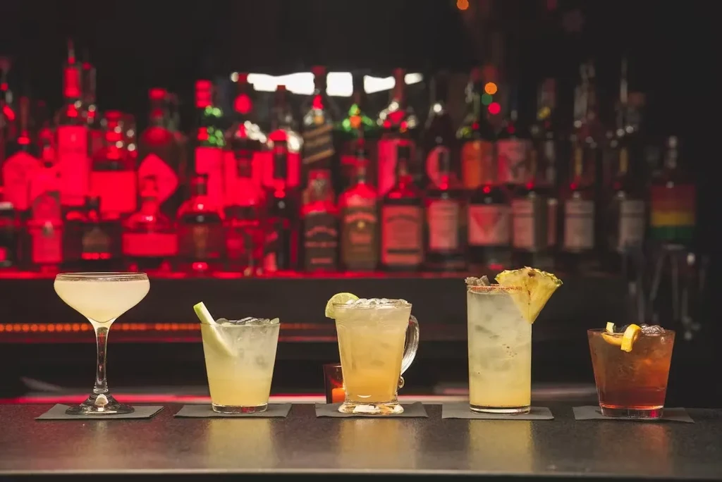 Five cocktails on a bar counter with bottles lit by red light.
