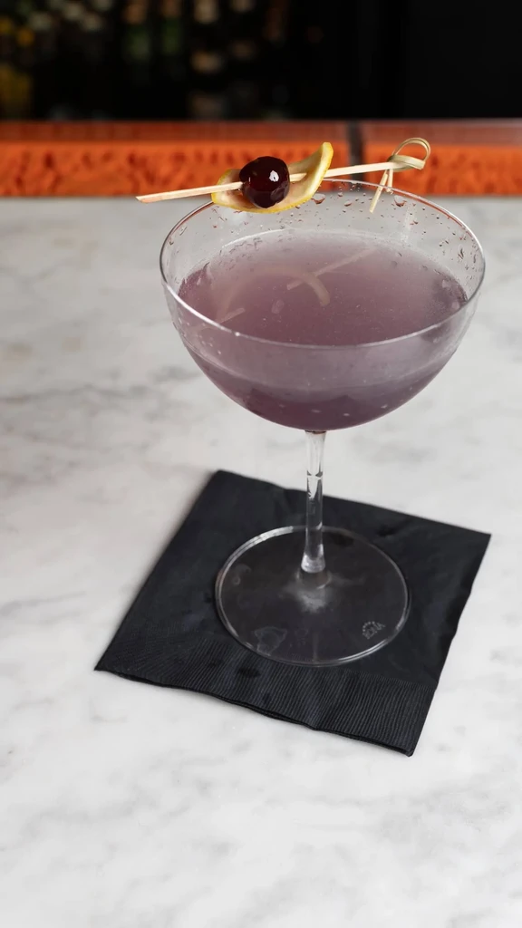 A purple cocktail in a coupe glass with a cherry-lemon garnish.