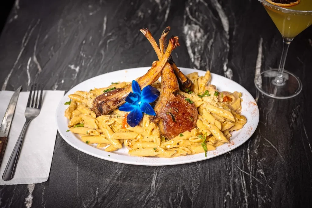 Lamb chops and penne pasta with a blue orchid garnish and cocktail.
