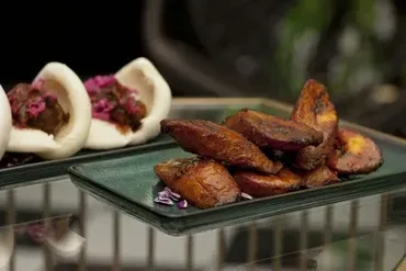 A plate of roasted plantains with meat-filled bao buns in the background.