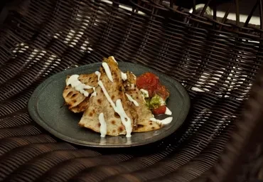Three quesadilla wedges with sauces on a dark plate within a wicker basket.