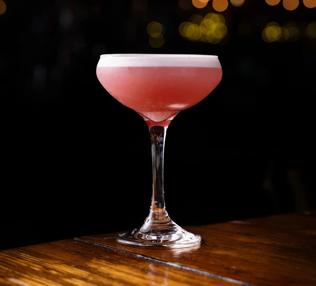 Pink cocktail with white foam in a coupe glass on a wooden table.