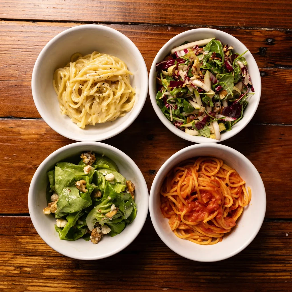 Four white bowls of pasta and salad on a wooden table.