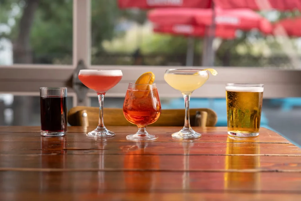Five alcoholic drinks lined up on a reflective wooden table.
