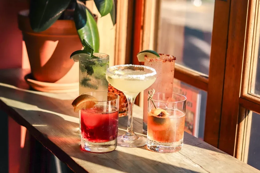 Five colorful cocktails on a wooden window sill in sunlight.