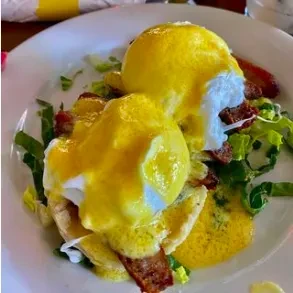 Two eggs Benedict with hollandaise sauce, bacon, and greens on a white plate.