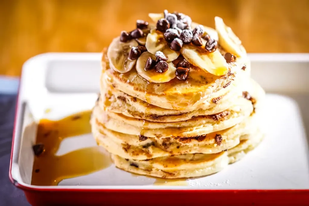 Stack of pancakes with bananas, chocolate chips, syrup, and powdered sugar.