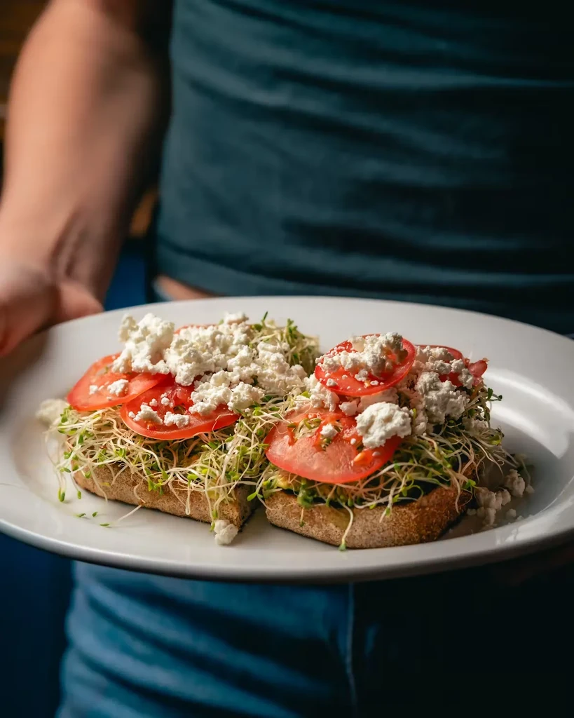 Two slices of toast with sprouts, tomatoes, and feta cheese.