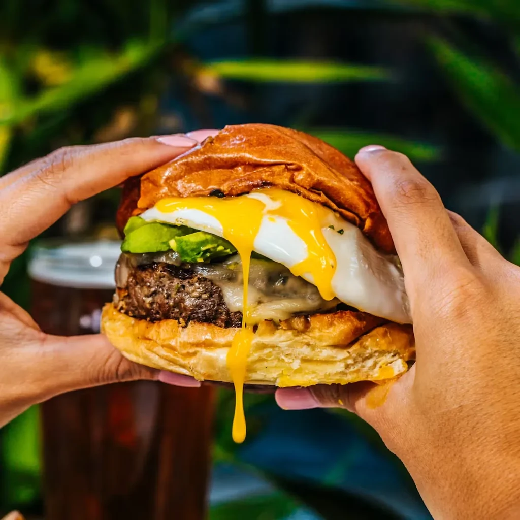 Hands holding a burger with melted cheese, avocado, and runny egg yolk.