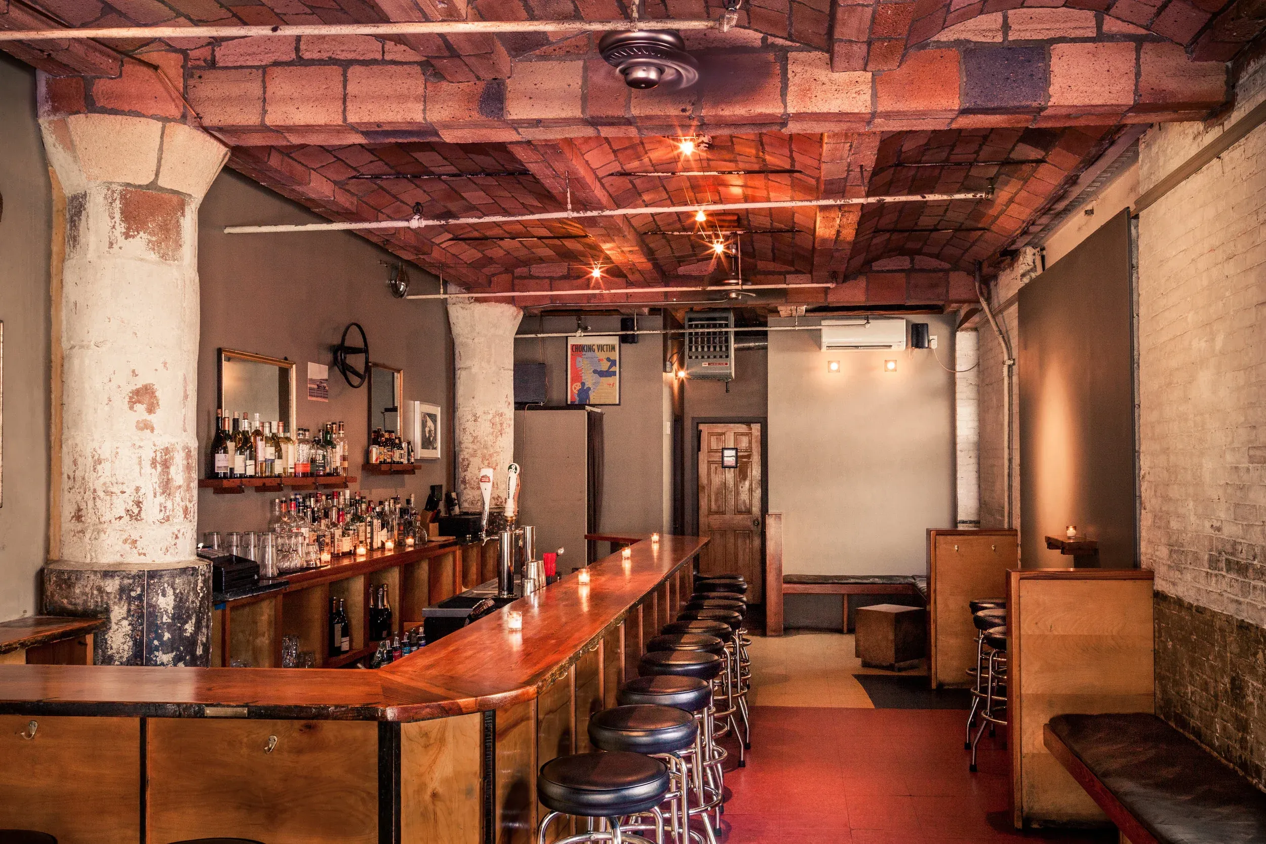 Rustic bar with a long wooden counter, stools, brick ceiling, and a red floor.