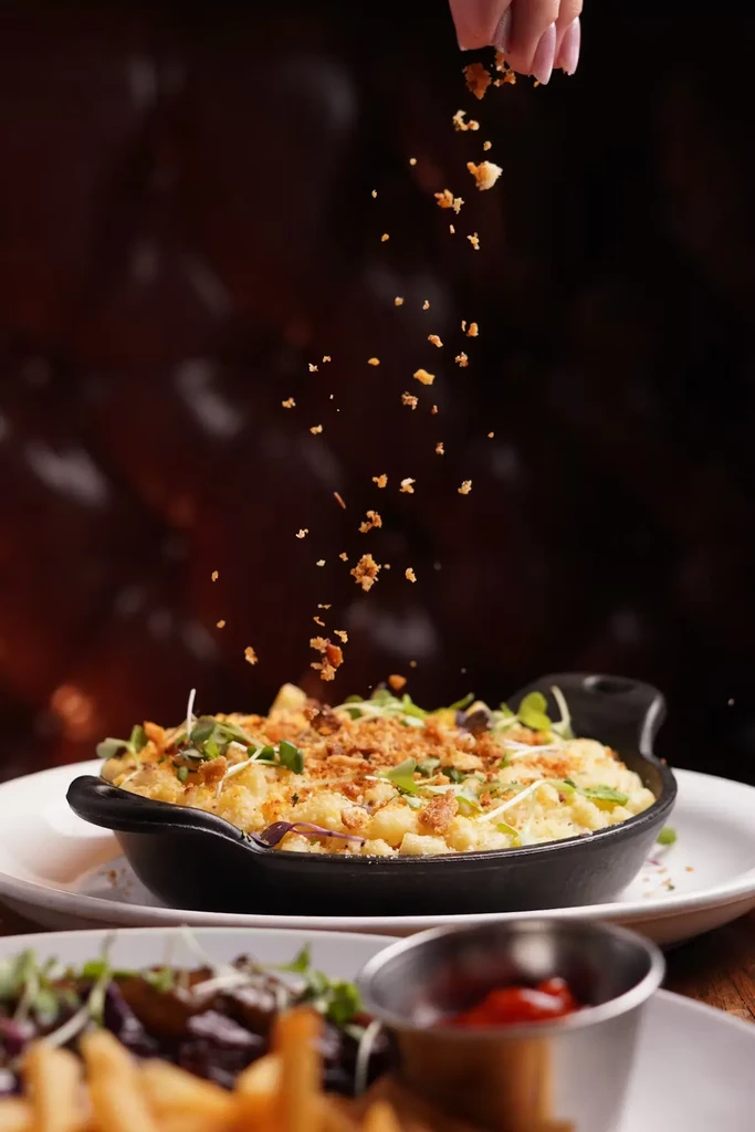 A hand sprinkles breadcrumbs over mac and cheese in a black bowl.