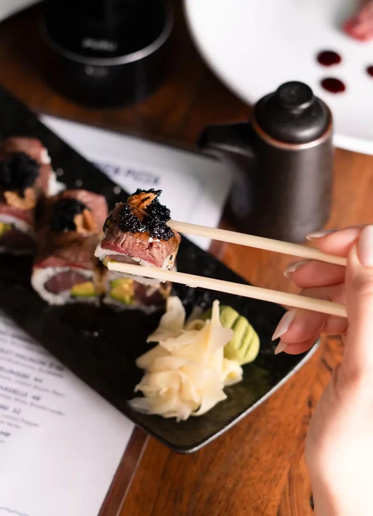Chopsticks holding a piece of sushi topped with steak and caviar.