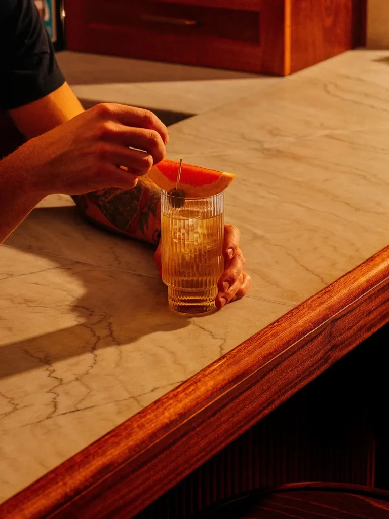 Hands garnishing an iced drink with grapefruit and olives on a bar.
