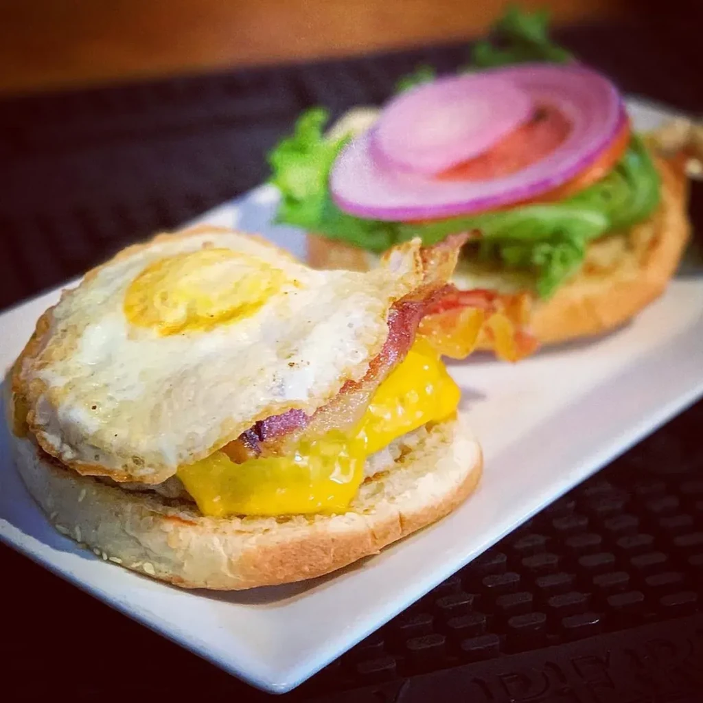 Two open-faced breakfast sandwiches with egg, bacon, cheese, lettuce, onion.