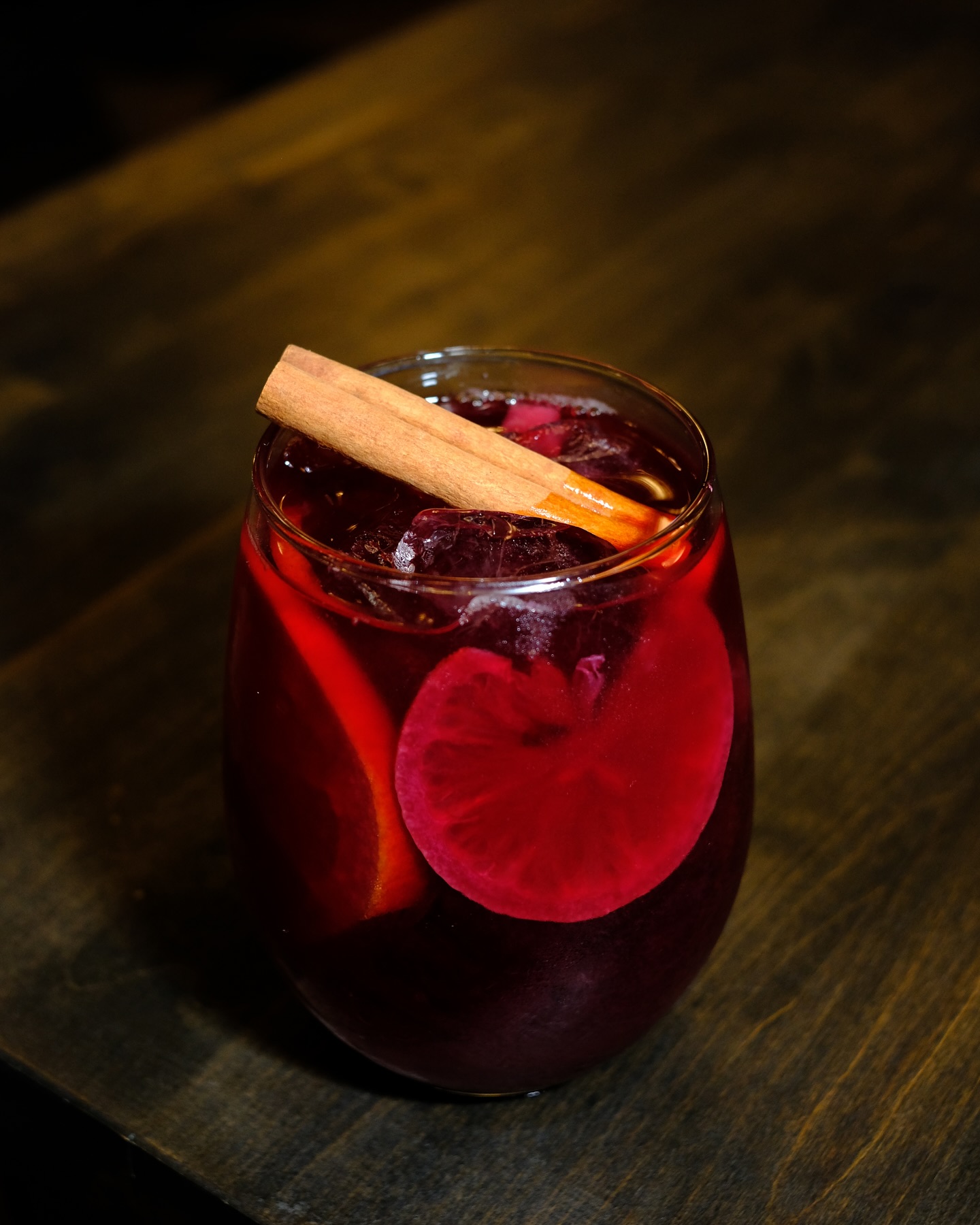 Glass of dark red drink with fruit and cinnamon stick.